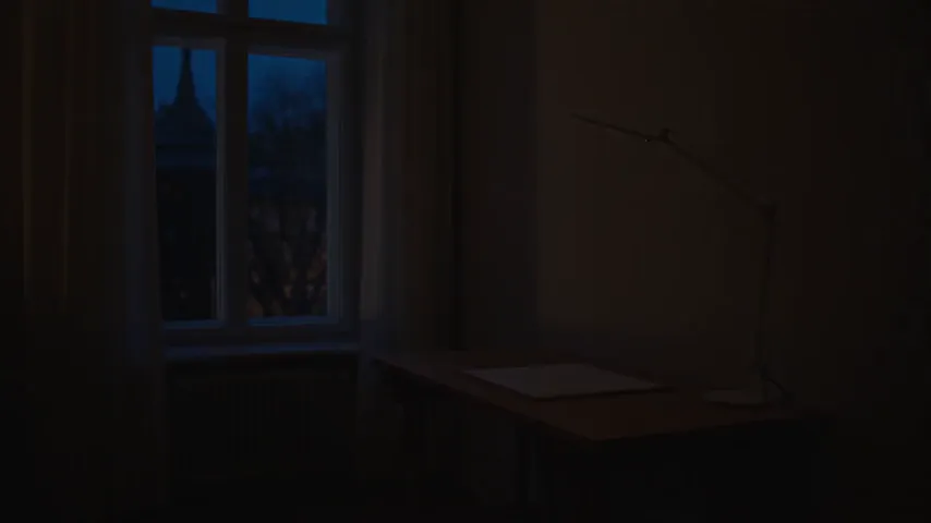 Seedance 2.5 test render of the desk lamp prompt: a designer adjusts a white desk lamp over a bare table, then the clip jumps to a close-up of the lamp arm in front of a pinned building sketch, with a blurred hand in the foreground.